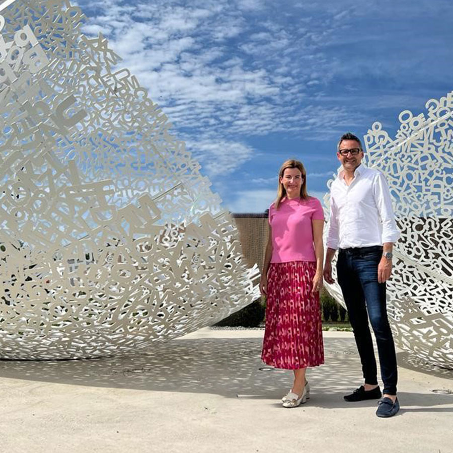 Eine Frau und ein Mann stehen lächelnd vor einer großen weißen Metallskulptur mit Buchstaben unter einem blauen Himmel.