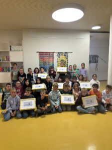 Eine große Gruppe von Grundschülern, die in einem Klassenzimmer Urkunden und gelbe Mappen in den Händen halten.