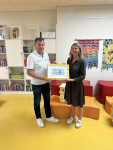 Ein Mann und eine Frau stehen gemeinsam vor den Regalen einer Bibliothek und halten ein gerahmtes Zertifikat in den Händen.