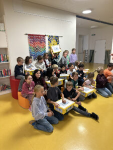 Eine Gruppe von Kindern sitzt auf dem Boden in einer Bibliothek; einige halten gelbe Plastikschachteln mit Urkunden in den Händen.