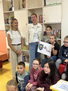 Zwei Frauen und sechs Kinder in einer Bibliothek; eines der Kinder hält ein Schild mit einem Logo und dem Wort Hubers in der Hand.