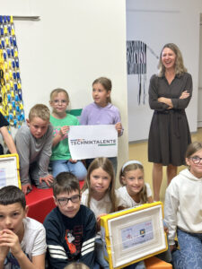 Eine Gruppe von Kindern hat sich um ein Schild mit der Aufschrift TECHNIKTALENTE versammelt; daneben steht eine lächelnde Frau.